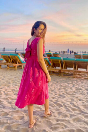 Dr.Elakshi Gupta More wearing a Strawberry Pink handloom Ikat midi dress with two pockets, standing on a sandy beach during sunset; sustainable cotton fashion by Abravi.