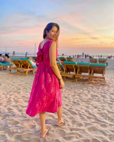 Dr.Elakshi Gupta More wearing a Strawberry Pink handloom Ikat midi dress with two pockets, standing on a sandy beach during sunset; sustainable cotton fashion by Abravi.